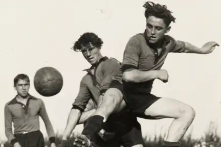 Une photographie en noir et blanc d'un match de football. L'un des joueurs se tient en l'air alors que sa jambe vient de taper dans le ballon.