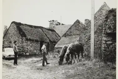 Une vue de batiment de ferme avec des chevaux au premier plan et deux hommes à leur côté