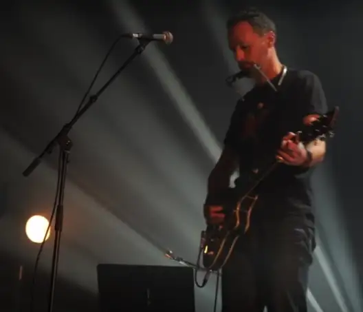 Un homme est à la guitare sur une scène devant un micro. La lumière des projecteurs filtre derrière lui.