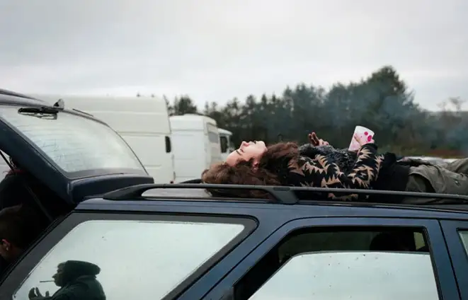 Sur le toit d'une voiture,  une jeune fille en manteau à fourrure est allongée.