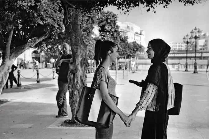 Sur une grande place devant un arbre, deux jeunes femmes se tiennent la main, l'une voilée et l'autre non.