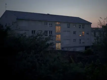 Une photo d'un petit immeuble à la tombée de la nuit. La cage de l'ecalier est allumée. Il y a également de la lumière dans l'un des appartements.