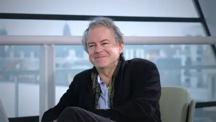 Yannick Haenel est assi dans un fauteuil, devant les vitres de la Bibliothèque, et souris à la personne qui lui parle. Il porte une veste en velours noir, une chemise bleue pâla et un foulard noir et or.