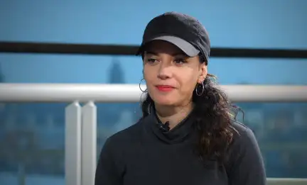 Cécile Adjadi est assi dans un fauteuil, devant les vitres de la Bibliothèque, et souris à la personne qui lui parle. Elle porte une casquette et un rouge à lêvres vif..