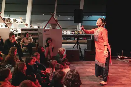 Sur cette image, une femme en tenue traditionnelle indienne fait une démonstration de danse dans la Bibliothèque des enfants. Devant elle, un groupe d'enfants observe attentivement sa performance.