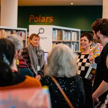 Dans les rayons de la Bibliothèque, une autrice parle en souriant à une assemblée de personnes debout.